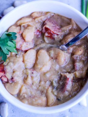 White bowl of cooked butter beans with sliced smoked sausage garnished with parsley leaves in bowl.
