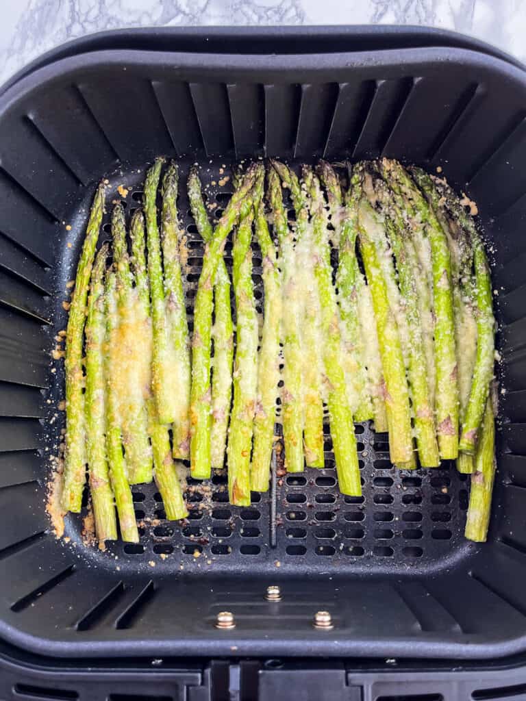 Fully cooked asparagus spears in air fry basket covered in melted parmesan cheese.