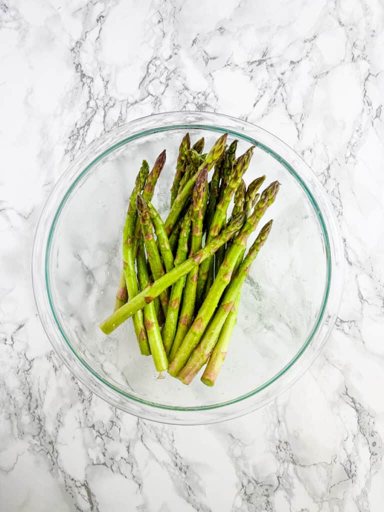 Uncooked asparagus in clear bowl tossed with olive oil and seasonings.