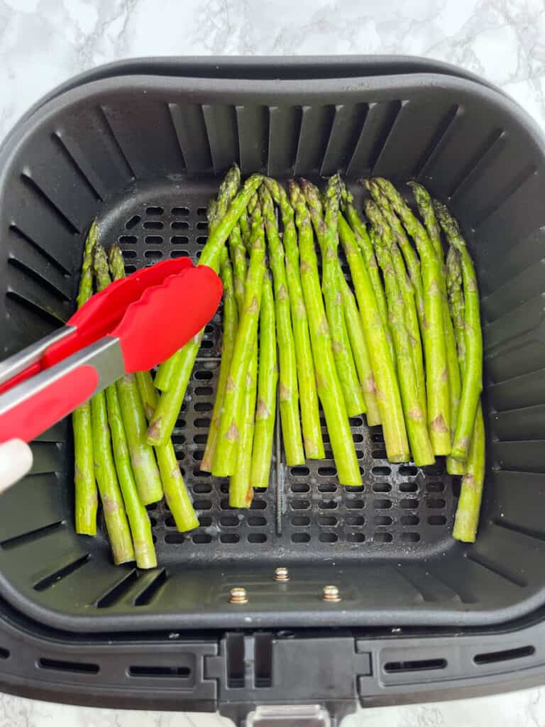 Uncooked asparagus being place in air fryer basket with red tongs.