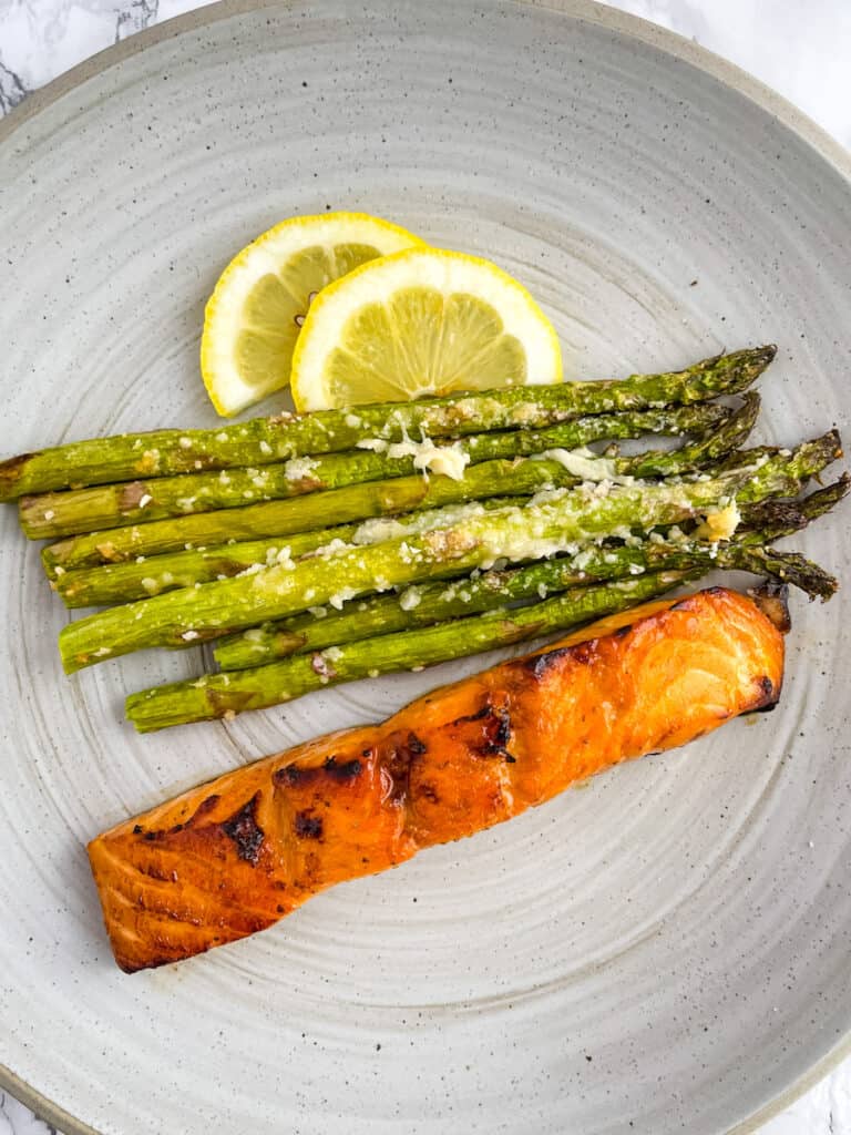 Salmon filet on a plate next to asparagus spears topped with melted parmesan cheese, garnished with lemon wedges.