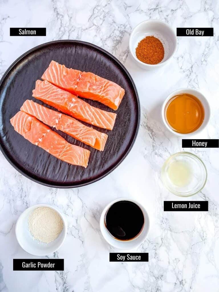 Five separate small ingredient bowls containing garlic powder, soy sauce, lemon juice, honey and old bay seasoning surrounding a plate containing 4 fresh salmon filets.