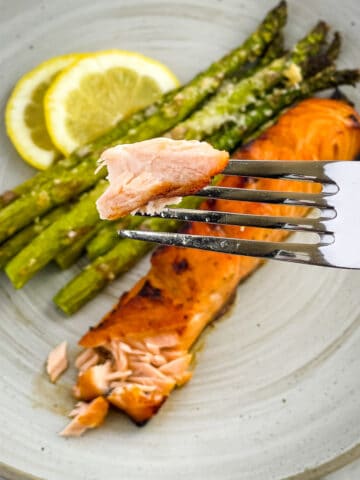 Piece of cooked salmon on fork in forefront and a plate containing the salmon filet and asparagus in the background.
